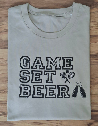 Flach zusammengelegtes Herren-T-Shirt in Beige mit dem Aufdruck „GAME SET BEER“ auf Holzoberfläche.