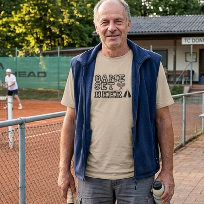 Ein etwa 50-顕hriger Mann steht lächelnd auf einer Tennisanlage und trägt ein beiges T-Shirt mit der Aufschrift 'Game Set Beer'. In der Hand hält er einen Tennisschläger.